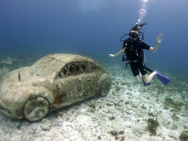 A sculpted car resting on the sand, a diver waving beside it